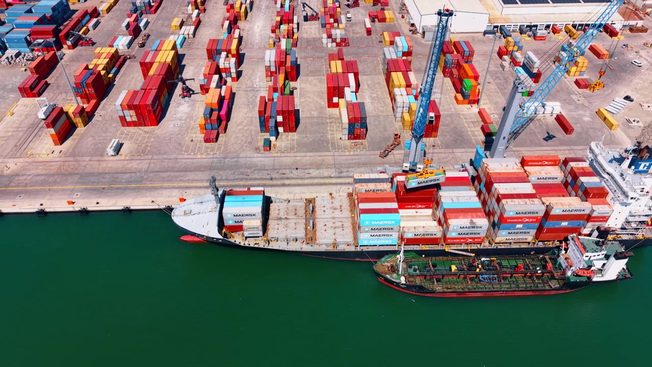 Burgas, Bulgaria, 1 May 2026: Top aerial view of container ship and quay. High angle drone view of container ship, service vessel, quay cranes, container yard and green harbor water.