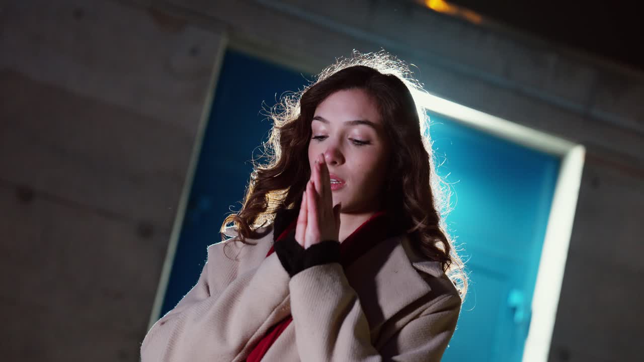 Ripresa notturna cinematografica di una donna nel freddo invernale con il respiro visibile mentre espira, che si scalda le mani sotto una forte retroilluminazione con bagliori dell'obiettivo. Concetto di clima gelido, solitudine.