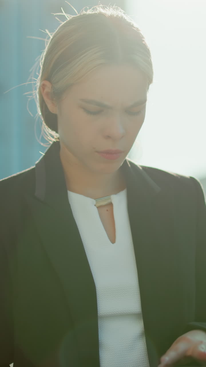 Bank manager dressed in formal outfit walking outdoors reviewing paperwork in folder under warm sunlight with slight lens flare and urban backdrop featuring glass buildings and parked cars