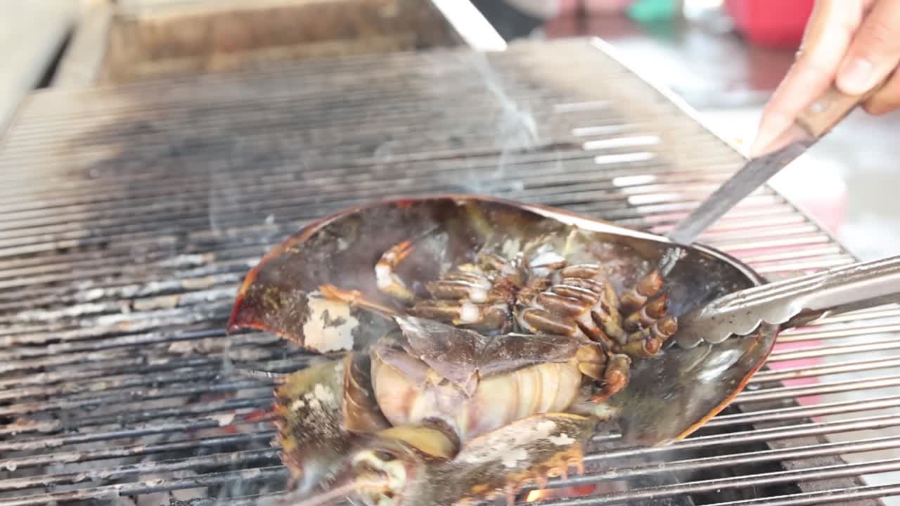A vendor uses tongs and a knife to cook a horseshoe crab over a grill