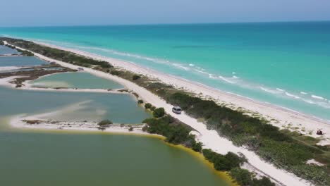 panoramic drone shot of camper van driving white sandy road between turquoise caribbean ocean and green salt lake view. mexico, rio lagartos,