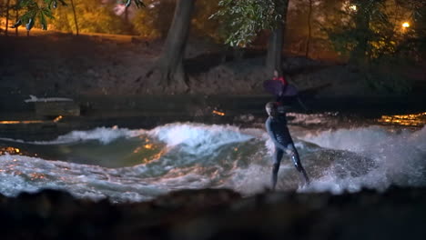 Cinematográfica-Cámara-Lenta-Eisbach-Ola-Río-Comunidad-De-Surfistas-Munich-Alemania-A-Través-De-árboles-Nivel-Del-Suelo-Caer-Otoño-Hermosa-Noche-Surf-Alto-Flujo-Agua-Surf-Alta-Baviera-Alpes-Fluir-Seguir-Movimiento