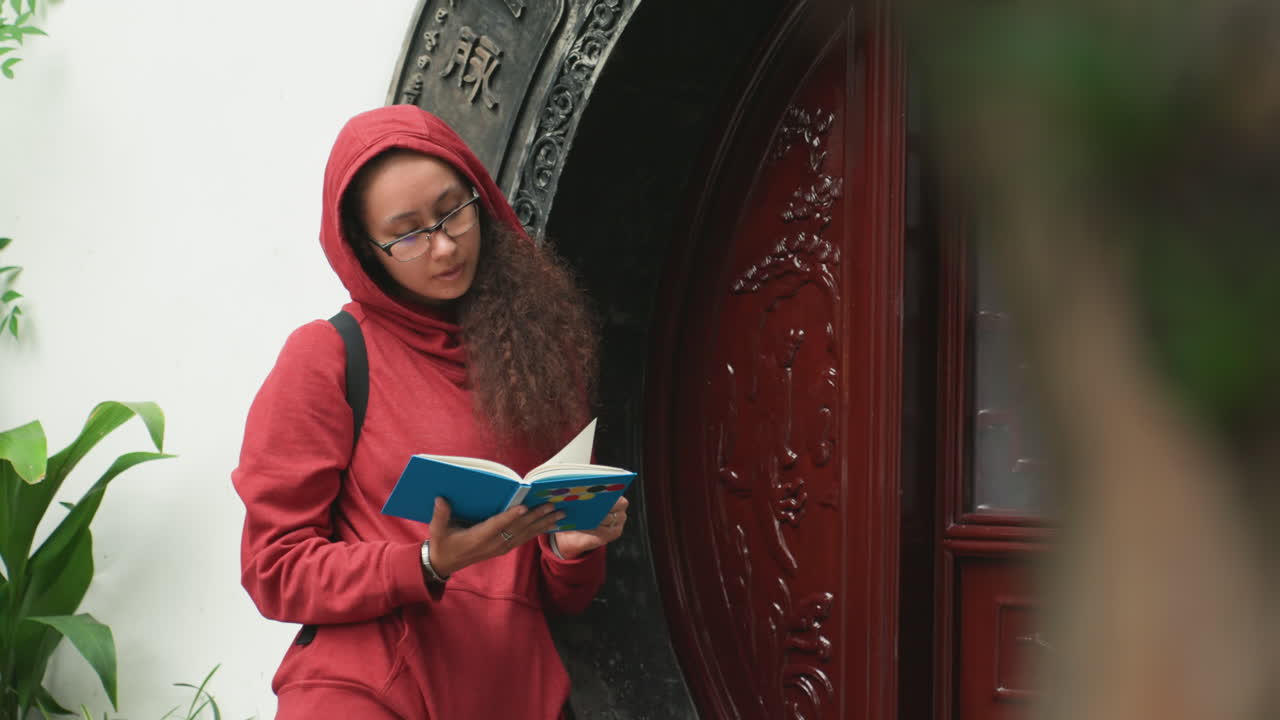 Wanita muda membaca buku di dekat ambang pintu berukir, mengenakan jaket merah berkerudung, ransel, gerimis, dedaunan hijau, pintu masuk kayu bundar berukir, kontemplasi yang tenang, cahaya lembut yang menyebar, senyum lembut, taman kota.