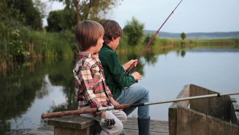 side view of two teen boys sitting on the lake pier, talking and fishing together