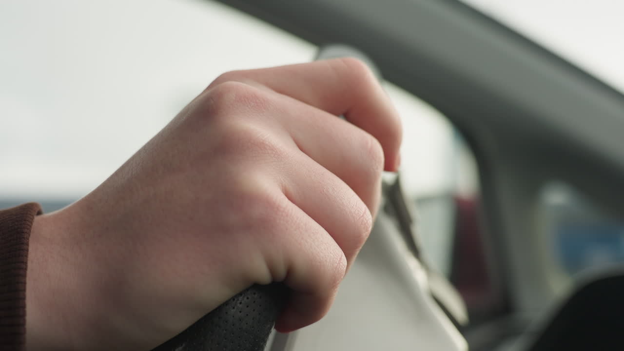 close up of driver hand holding steering wheel with firm grip while slightly tapping surface, showing control and focus during drive with soft lighting and blurred window view in background