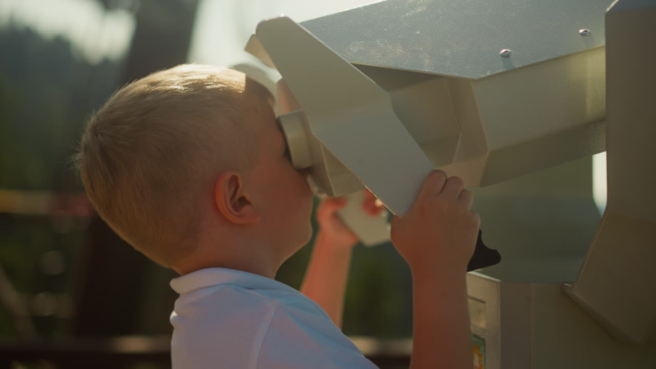 niño inquisitivo explora el mundo a través de binoculares masivos. niño pequeño en polo blanco disfruta de la maravillosa vista de las montañas onduladas de pie en la cubierta de observación