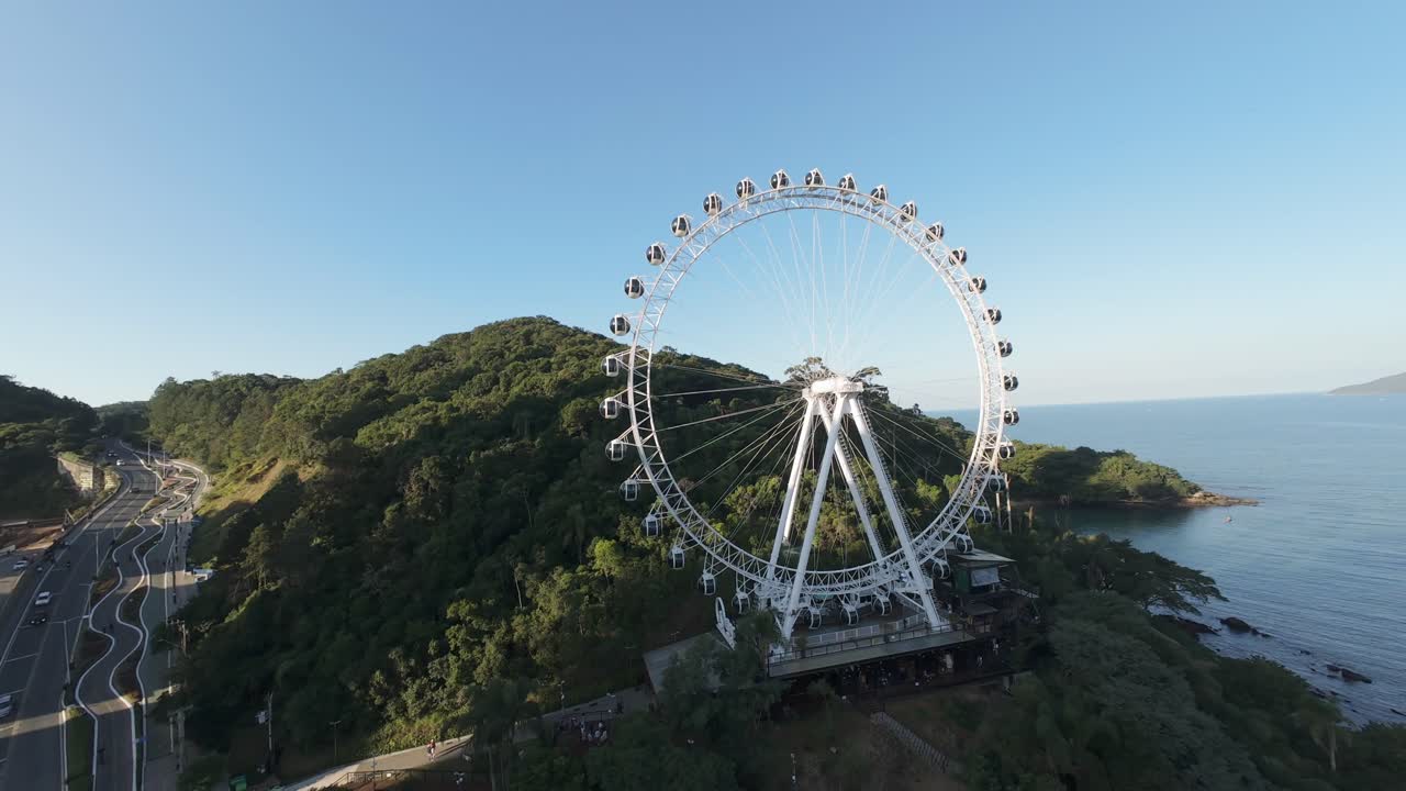 FPV drone performs extreme wide-angle turn around the iconic Ferris wheel of Balneário Camboriú, Santa Catarina, Brazil, revealing lforest below and the Gulf of Balneário Camboriu