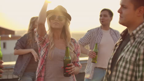 Una-Mujer-Con-Gafas-Y-Sombrero-Se-Mueve-Bailando-Con-Sus-Dos-Amigas-Y-Tres-Novios-En-La-Azotea.-Ella-Sonríe-Y-Disfruta-Del-Tiempo-Con-Una-Cerveza-En-Una-Tarde-De-Verano.