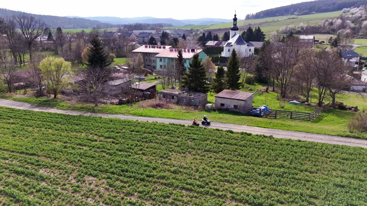 Vista aérea de un paisaje rural con campos y casas mientras dos niños montan quads por un camino, un paisaje tranquilo con colinas ondulantes y ambiente primaveral