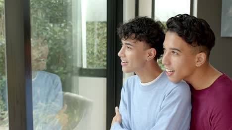 smiling multiracial gay couple looking out window, enjoying time at home