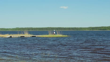 beautiful view of a lake usma shore on a sunny summer day, man fishing from the wooden pier, distant islands with lush green forest, rural landscape, coast with old reeds, distant wide shot