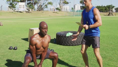 Two-diverse-fit-men-exercising-outdoors,-one-squat-lifting-kettlebells-while-the-other-times-him