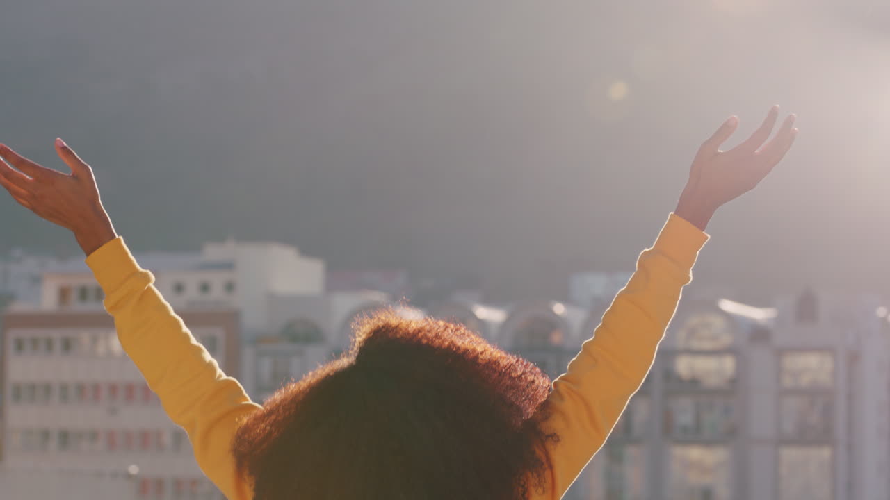 뒷면 행복한 아프리카계 미국인 여성 팔을 들고 지붕 위에서 자유를 축하하고 해가 지는 도시에서 독립적인 생활 방식을 즐기고 있습니다.