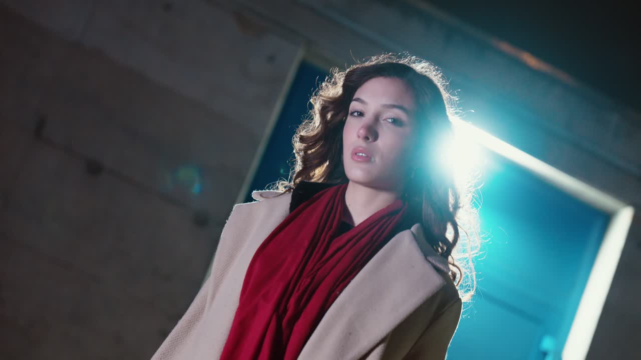 Ritratto notturno cinematografico di una bella donna in cappotto e sciarpa rossa, retroilluminata da una luce di sfondo brillante mentre il vento le muove i capelli. Concetto per moda urbana, atmosfera drammatica della vita notturna.