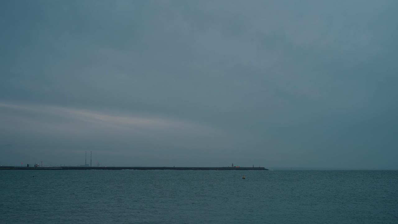 A long pier extends into the sea at Dún Laoghaire, with the Poolbeg chimneys visible in the background. The sky is overcast, and soft evening light is reflecting on the water.