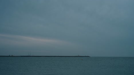 a long pier extends into the sea at dún laoghaire, with the poolbeg chimneys visible in the background. the sky is overcast, and soft evening light is reflecting on the water.