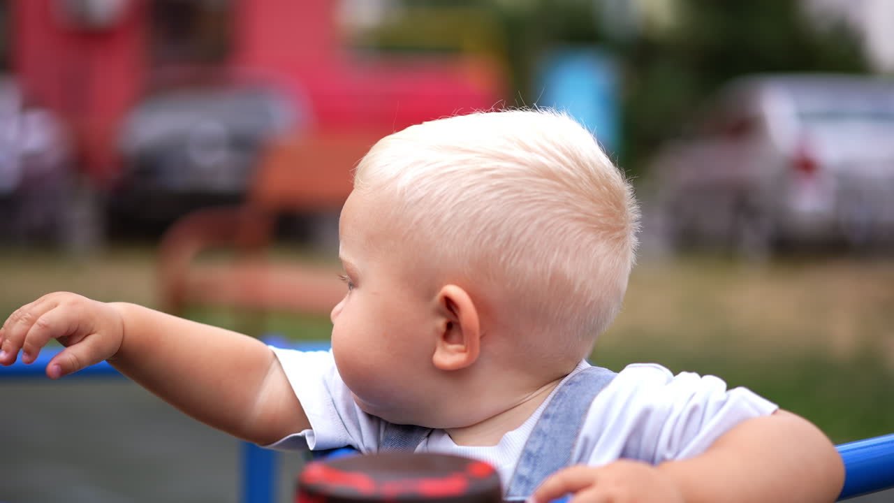 Adorable blond Caucasian toddler stands on the carousel. Portrait of a happy baby boy close up. Blurred backdrop.