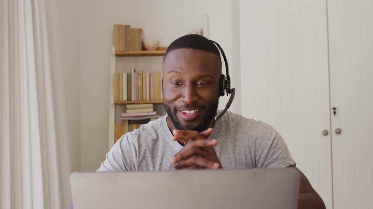 African american man using phone headset and having a video chat on laptop while working from home