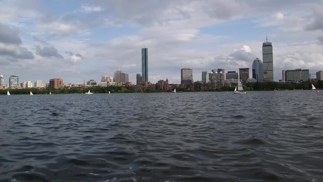 Skyline di Boston visto dal fiume Charles in una giornata parzialmente nuvolosa, con barche a vela sull'acqua e moderni grattacieli del centro in lontananza.