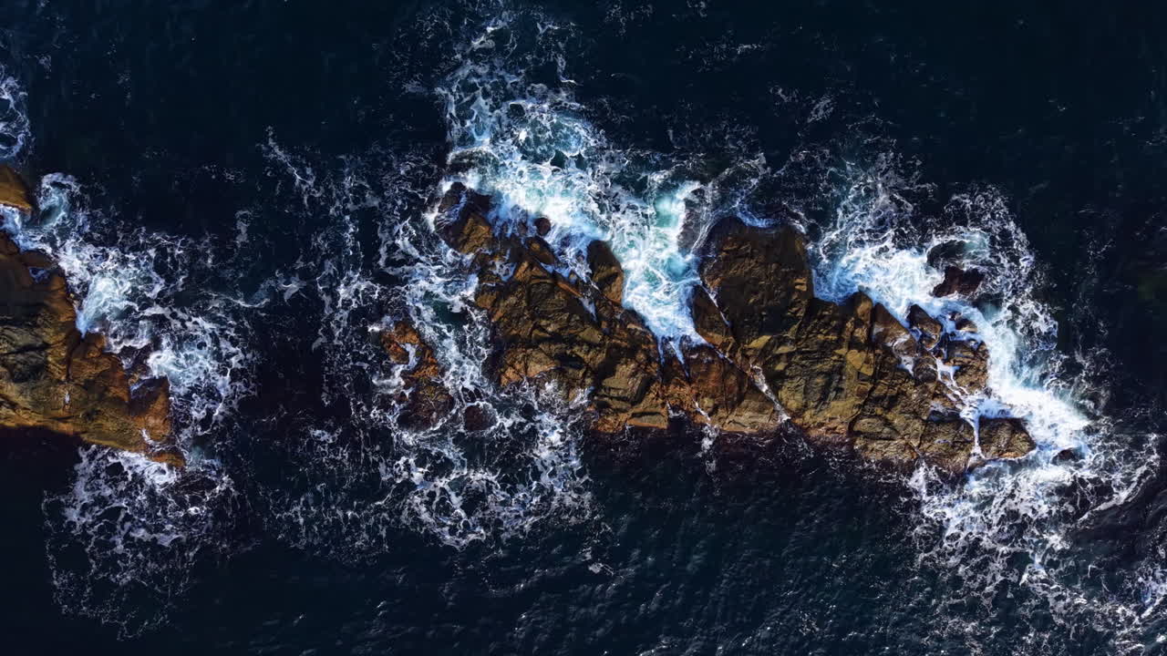 Onde che si infrangono contro scogliere aspre sotto un cielo azzurro limpido sulla costa rocciosa durante un pomeriggio estivo luminoso.