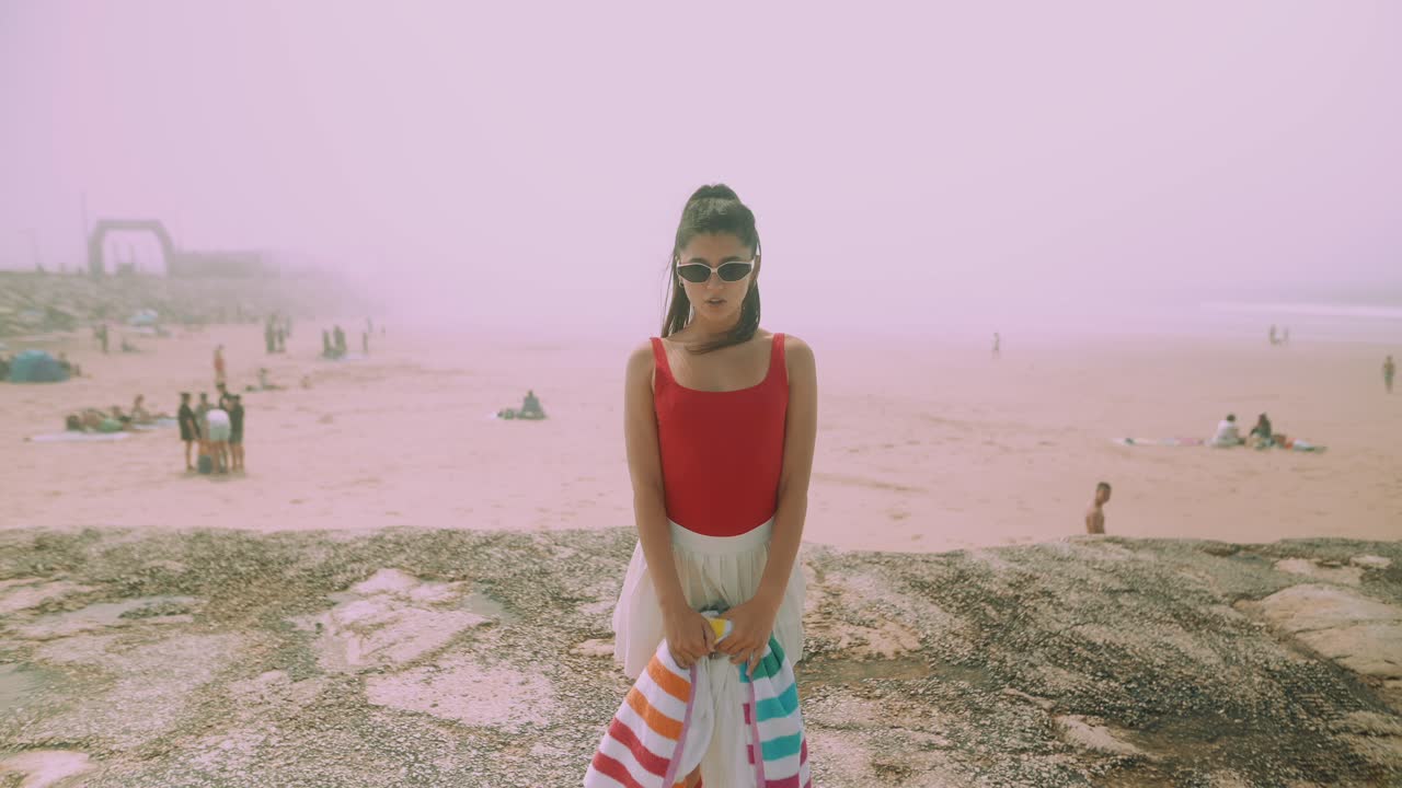 Giovane donna in costume da bagno rosso e occhiali da sole in piedi su una spiaggia nebbiosa