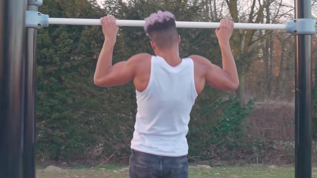 Panorering rundt om en muskuløs mand, der laver pull-ups i en calisthenics park ved solnedgang.