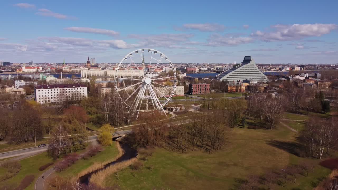 A zoom-out shot of the Ferris wheel in Pardaugava, Riga, revealing the stunning cityscape and business buildings under the warm sunshine.