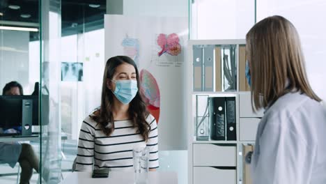 doctora caucásica con mascarilla médica sentada en el escritorio y explicando a una paciente el tratamiento del coronavirus en consulta médica