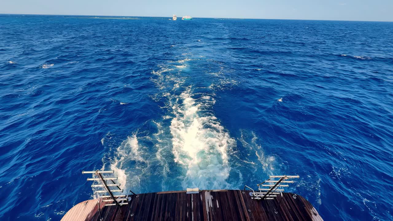 Eine ruhige Draufsicht auf das Teakholzdeck einer Luxusyacht, die an einem sonnigen Sommertag durch tiefblaues Mittelmeerwasser gleitet und eine lebendige weiße Gischtspur unter einem klaren Himmel erzeugt.