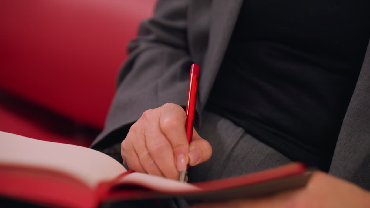 Close-up shot of person writing notes in notebook with red pen, business, productivity, and planning concept. Focus on hand holding pen and writing, blurred background in modern workspace