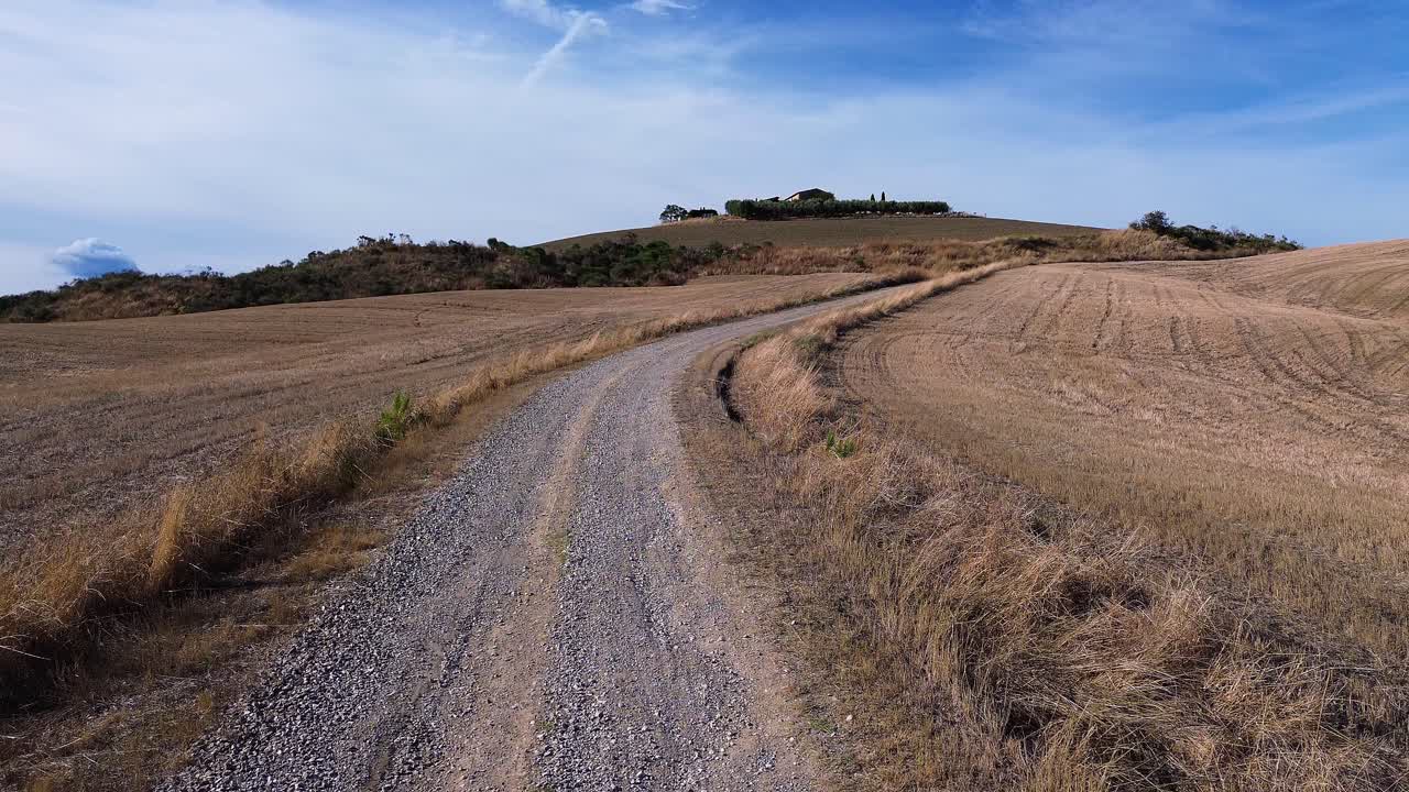 A winding gravel road through the golden fields of Tuscany, Italy, with rolling hills and a farmstead in the distance