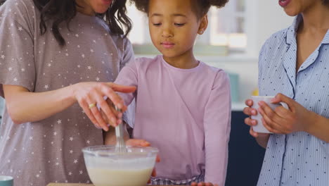 family with two mums wearing pyjamas making morning pancakes in kitchen at home with daughter