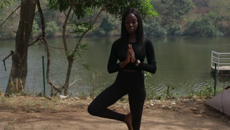 a lady during yoga in the tree pose by a lake, static wide shot