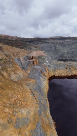 vertical fpv drone flight closely explores the mineral-stained, colorful textured walls of an open-pit mine, revealing geological strata and deep water. industrial, geological survey