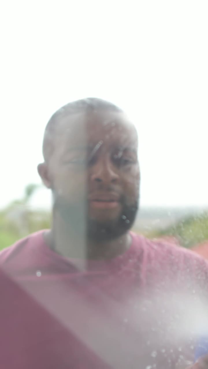 Vertical video: Raising arm, man in maroon tee wiping patio window with squeegee, clearing soap