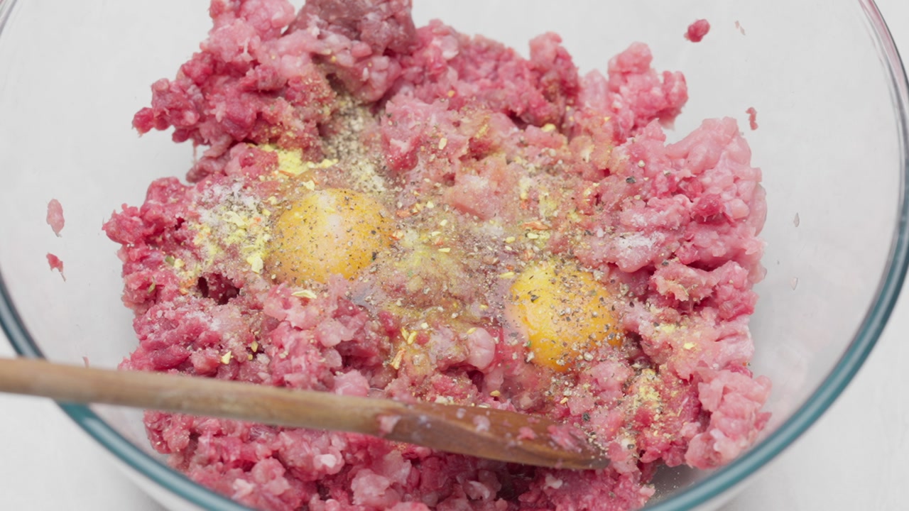 Overhead close-up of spices added to raw ground meat with egg in a bowl.