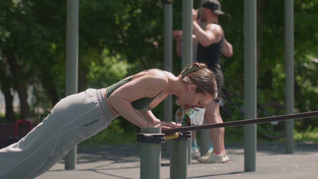Close up of young woman performing push ups on outdoor exercise bars with snack bag visible. Man in background exercises separately both surrounded by green park environment and sunlight