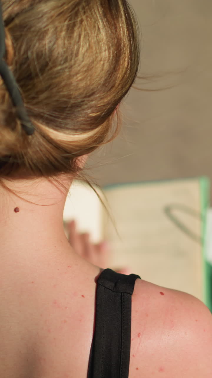 vista trasera de una mujer sentada en un banco al aire libre con una mancha en la espalda, recuperando un libro de una bolsa verde menta, abre el libro y hojea sus páginas mientras las sombras se proyectan en el suelo