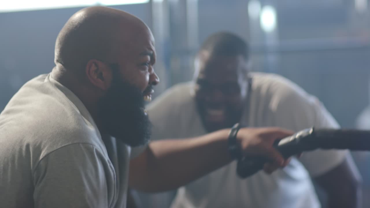 African American men training, coach leaning in encouraging, trainee pulling battle rope powerfully