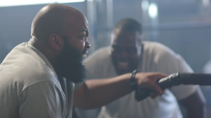 African American men training, coach leaning in encouraging, trainee pulling battle rope powerfully