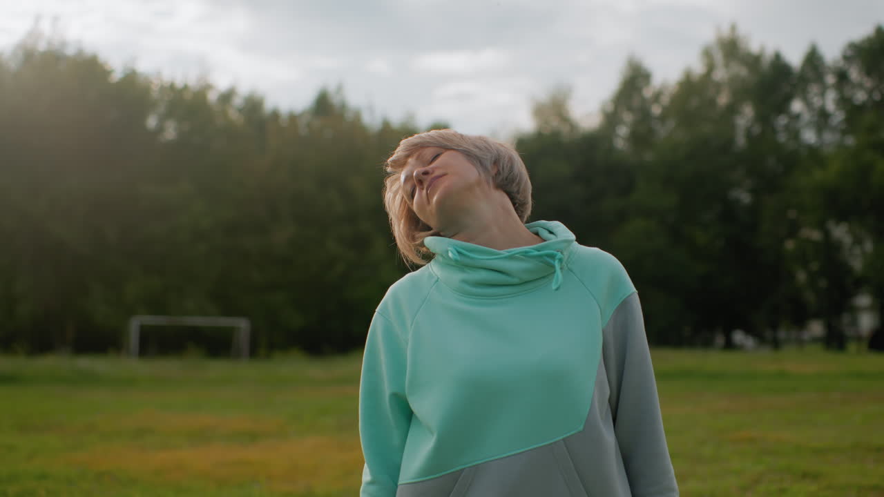 Trainer in mint tracksuit doing slow neck rotation exercise on open green field during peaceful morning workout session focusing on flexibility stretching relaxation posture correction
