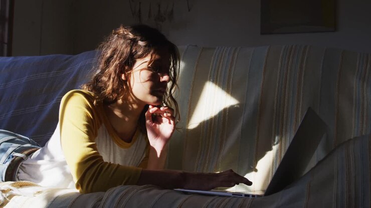 Donna di etnia mista felice, sdraiata sul divano, che usa il portatile e sorride nel luminoso soggiorno.