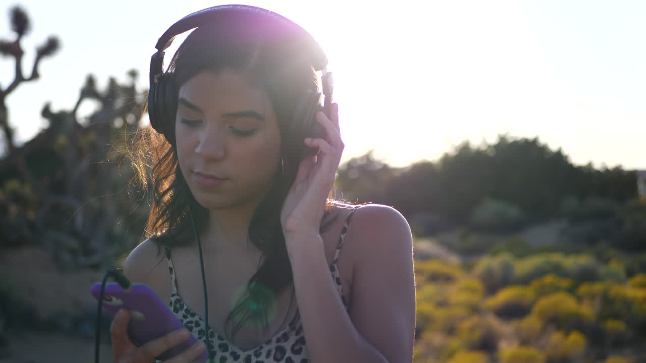 una hermosa joven hispana sosteniendo un teléfono inteligente escuchando música triste o grave en auriculares al aire libre en la luz del sol las luces de las lentes en cámara lenta