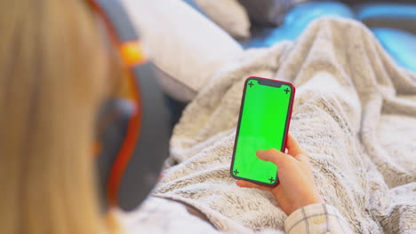 teenage girl on sofa wearing wireless headphones streaming content from green screen mobile phone