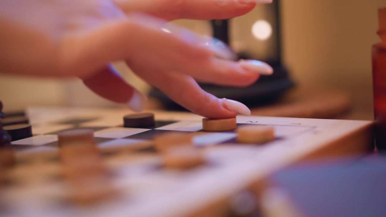 Primo piano di una mano con unghie lunghe e bianche che muove i pezzi del gioco su una scacchiera da dama. La scena presenta un'illuminazione soffusa e una profondità di campo ridotta, concentrandosi unicamente sull'interazione tra le dita e i pezzi di legno.