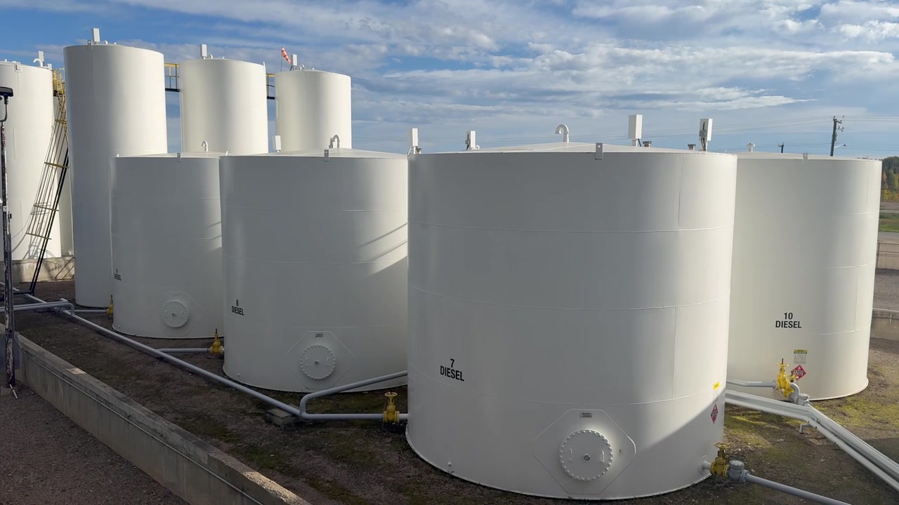 Um movimento de câmera panorâmica para a direita com um zoom lento em uma planta de armazenamento de petróleo em funcionamento, exibindo grandes tanques de armazenamento brancos e tubulações complexas em operação sob um céu parcialmente nublado