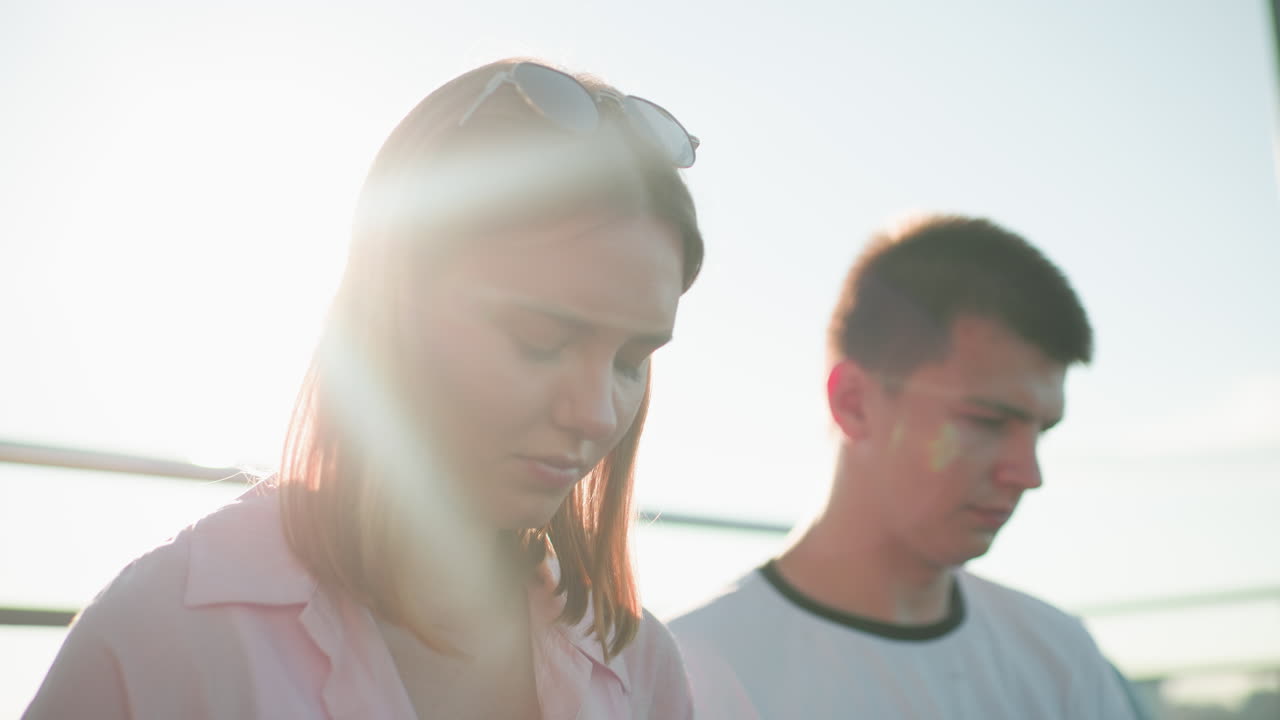 머리에 안경을 쓴 분홍색 셔츠를 입은 젊은 여성, 빛에 목욕하여 홀로 효과를 만들어, 집중된 젊은 남자의 에 서 있으며, 둘 다 금속 레일링 배경에 맞춰 있습니다.