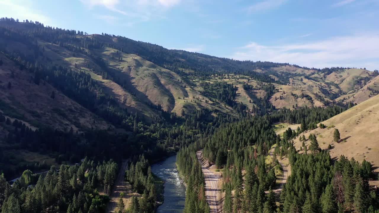 4K Drone Pushes in through a gully formed by a river. The river is lined with thick dense trees as the blue sky looks down on them. There is a road that follows the river as traffic flows.