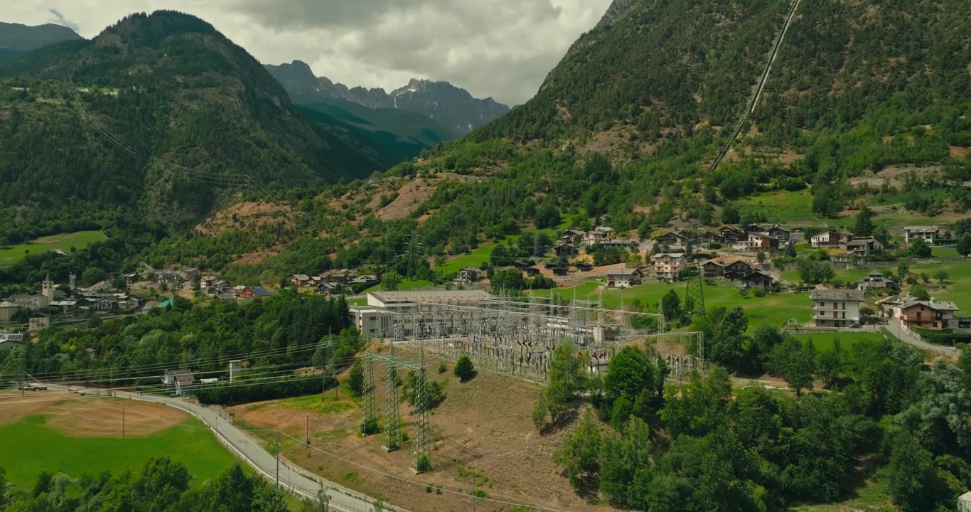 Il drone si alza sopra le cime degli alberi per rivelare una sottostazione elettrica dove convergono più linee di trasmissione in un pittoresco villaggio di montagna, mostrando l'infrastruttura energetica.