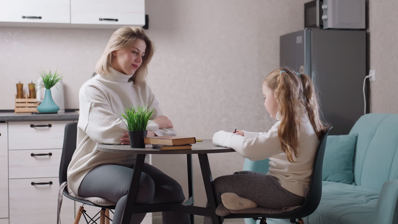 Kvinde sidder ved køkkenbord og interagerer med pige, der skriver i notesbog. Begge er iført hyggelige sweatre og omgivet af bøger og stueplanter, mens det moderne køkkeninteriør skaber en varm familiær atmosfære.